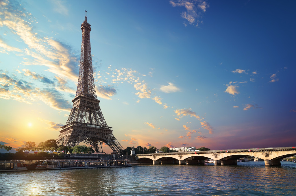 Eiffelturm in Paris bei Sonnenuntergang von der Seine aus gesehen. Eine beleuchtete Steinbogen-Brücke führt über den Fluss.