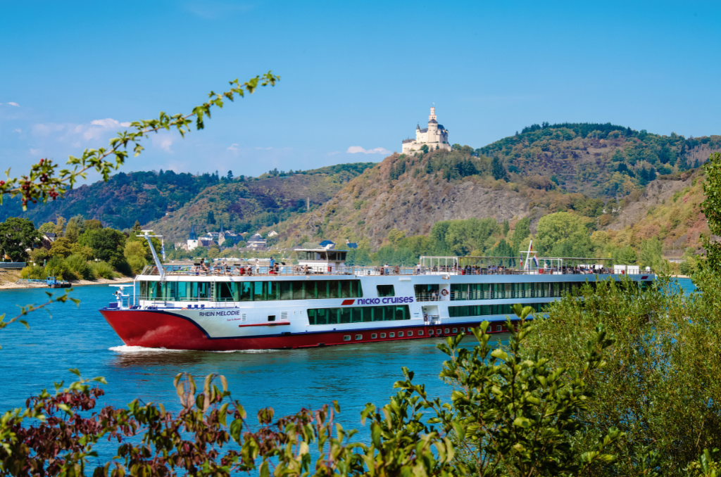 Das Flusskreuzfahrtschiff „Rhein Melodie“ von nicko cruises fährt auf einem Fluss vor einer hügeligen Landschaft mit einer Burg auf der Spitze.