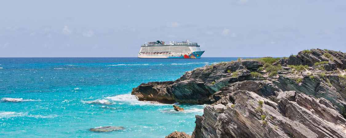 Gemini hat gesagt Ein großes Kreuzfahrtschiff mit bunter Rumpfbemalung fährt auf türkisblauem Meer an einer zerklüfteten Felsenküste vorbei.